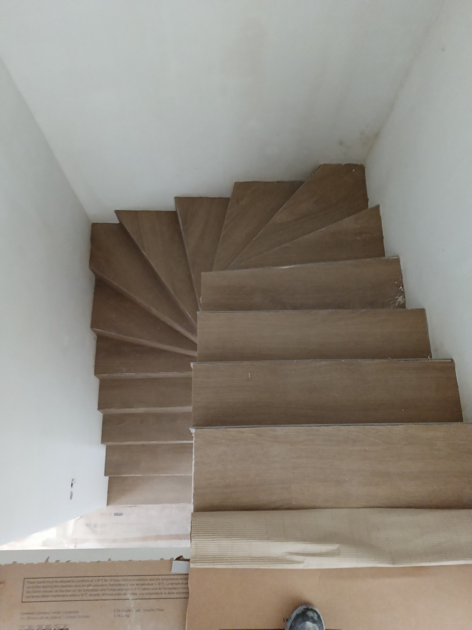 Spiralne schody wykończone panelami w odcieniach brązu, widok z góry. Częściowo widoczne opakowania paneli i fragment buta. Jasne ściany, surowy stan wykończenia.