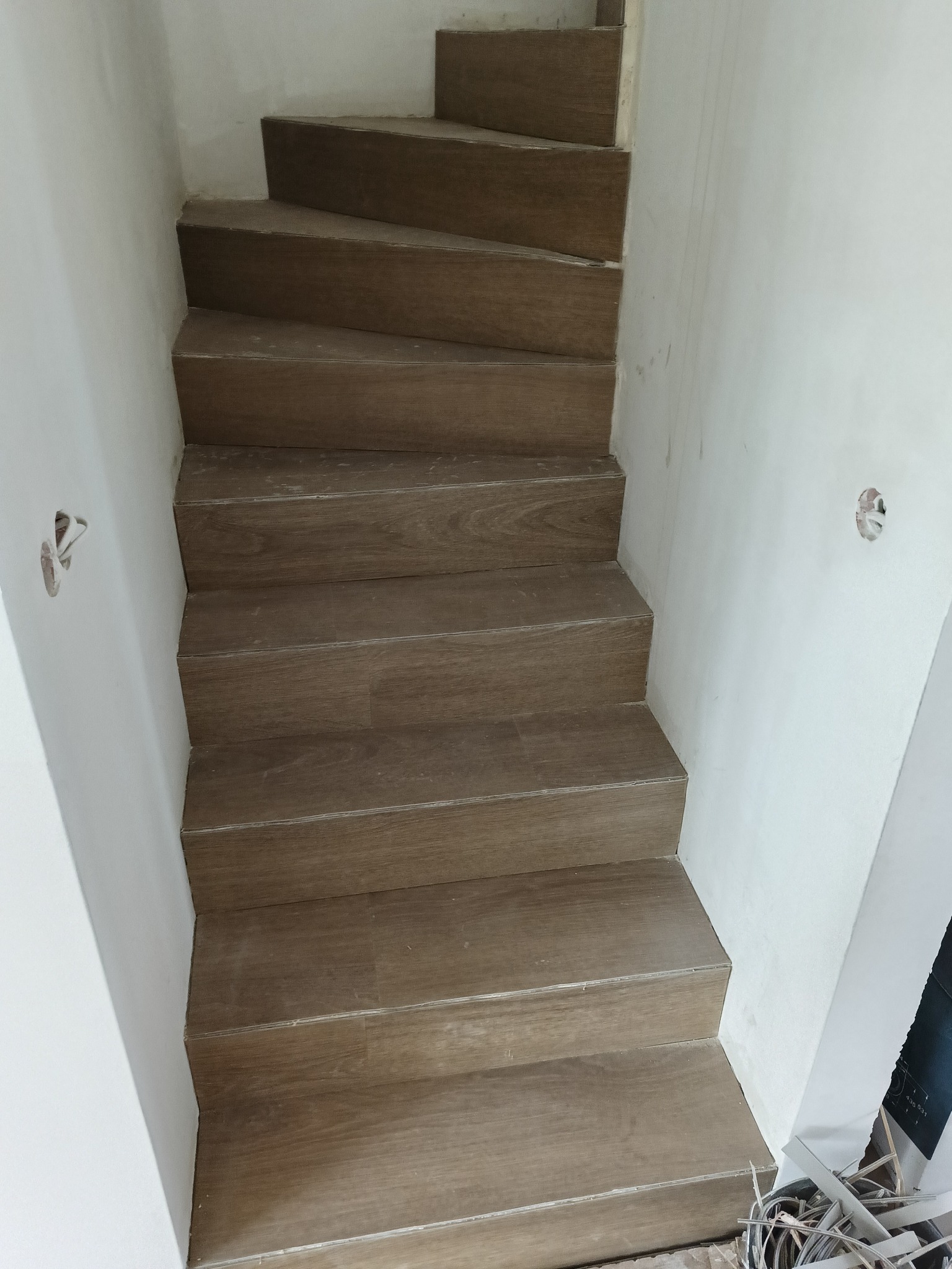 Wykończone schody z drewnopodobnych paneli, widok z góry. Białe ściany, widoczne puszki elektryczne. Ukończony montaż paneli na schodach.