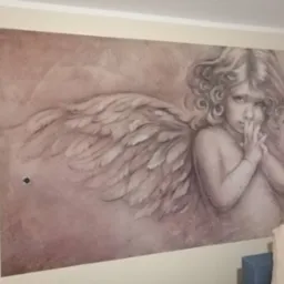 Malowidło ścienne przedstawiające anioła z kręconymi włosami i skrzydłami, stylizowane na sepię, z widocznym gniazdkiem elektrycznym po lewej stronie.