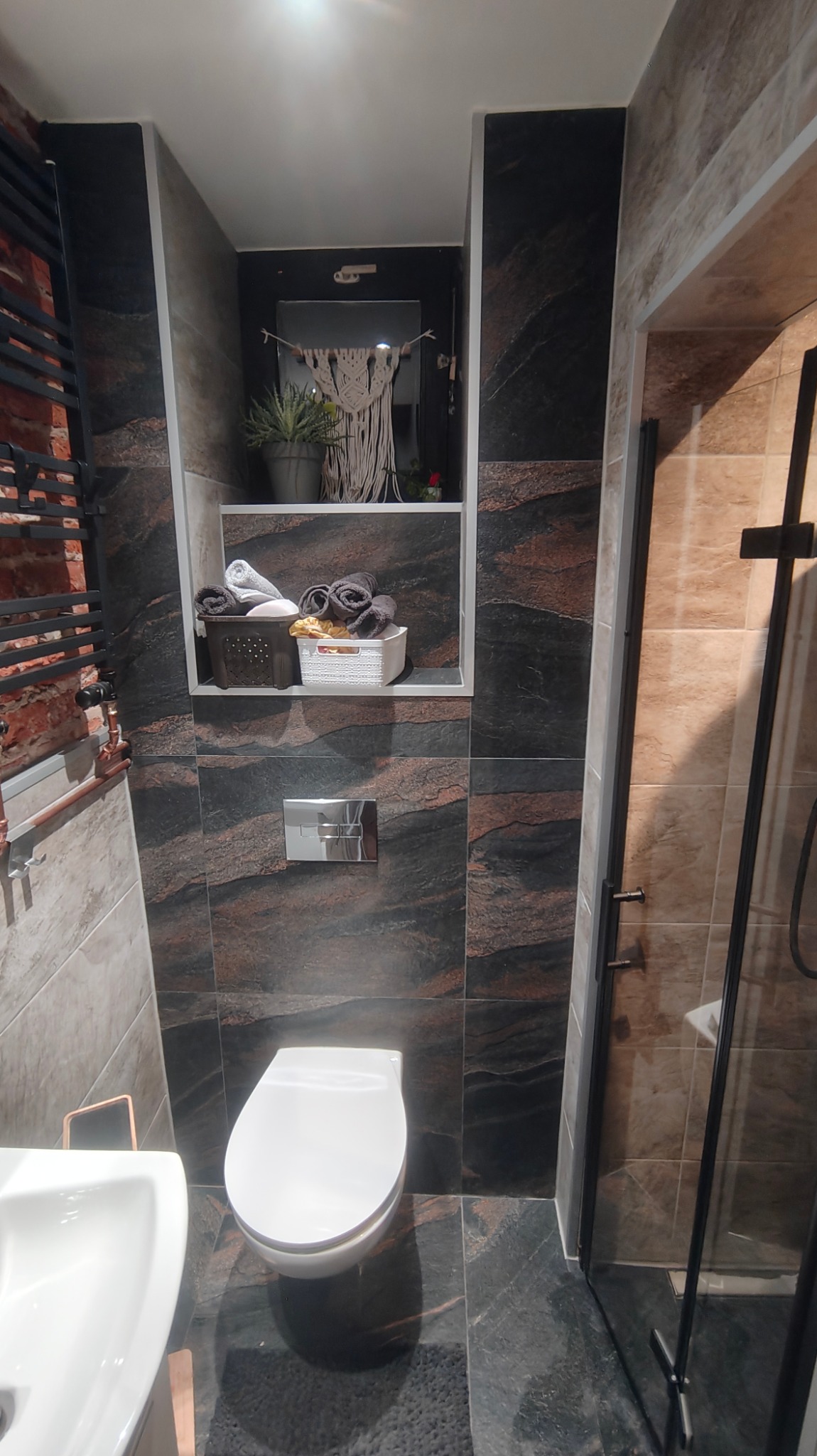 Nowoczesna łazienka z ciemnymi płytkami imitującymi kamień, podwieszana toaleta z chromowanym przyciskiem, umywalka z szafką w kolorze miedzi, wnęka z dekoracjami i ręcznikami, kabina prysznicowa...