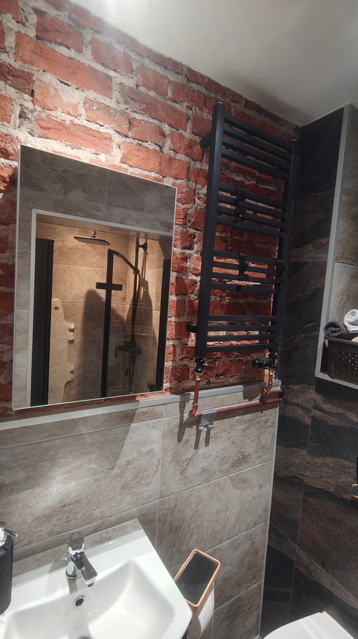 Nowoczesna łazienka z ceglaną ścianą, czarnym grzejnikiem drabinkowym z miedzianymi rurami, lustrem odbijającym kabinę prysznicową i umywalką.
