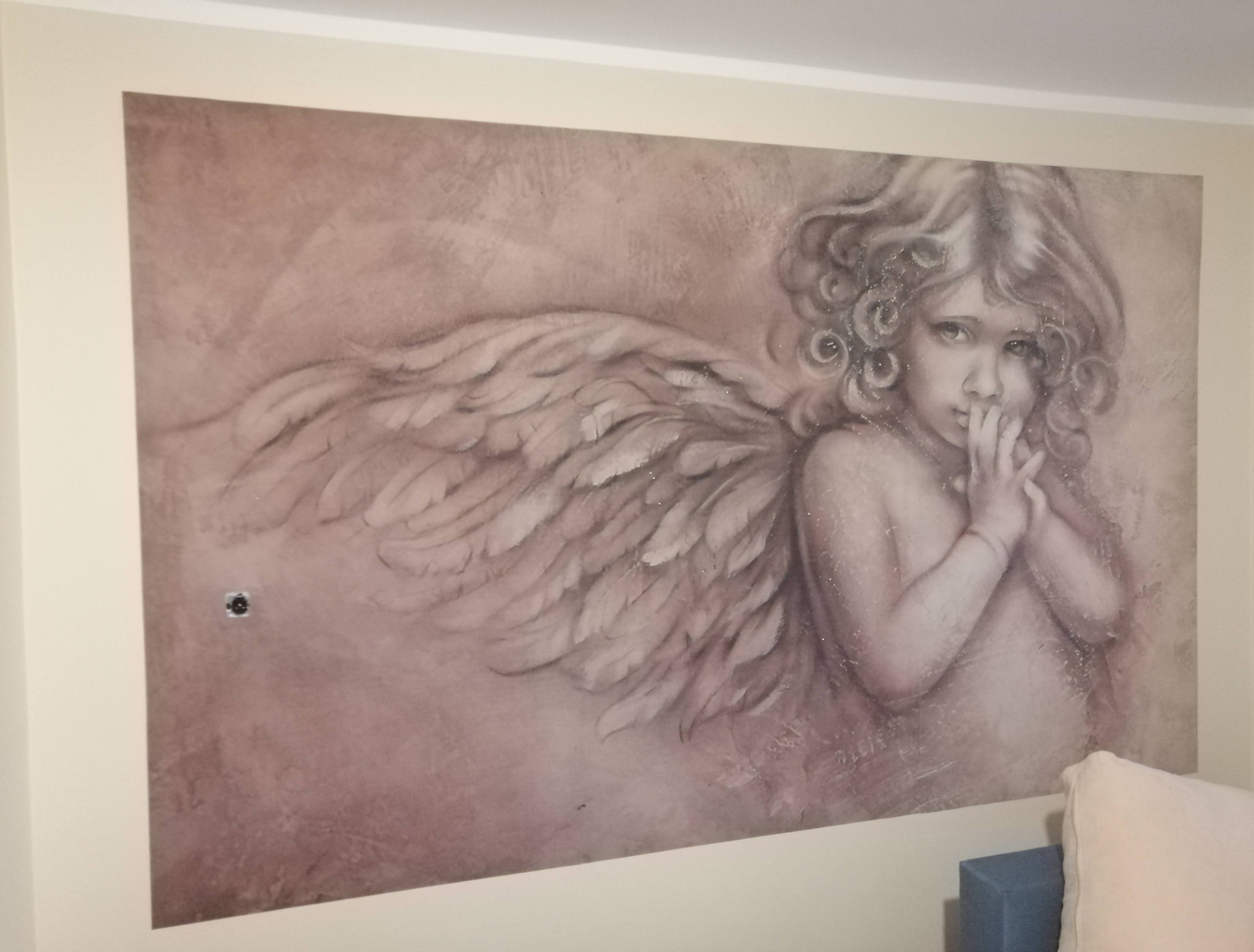 Malowidło ścienne przedstawiające anioła z kręconymi włosami i skrzydłami, stylizowane na sepię, z widocznym gniazdkiem elektrycznym po lewej stronie.