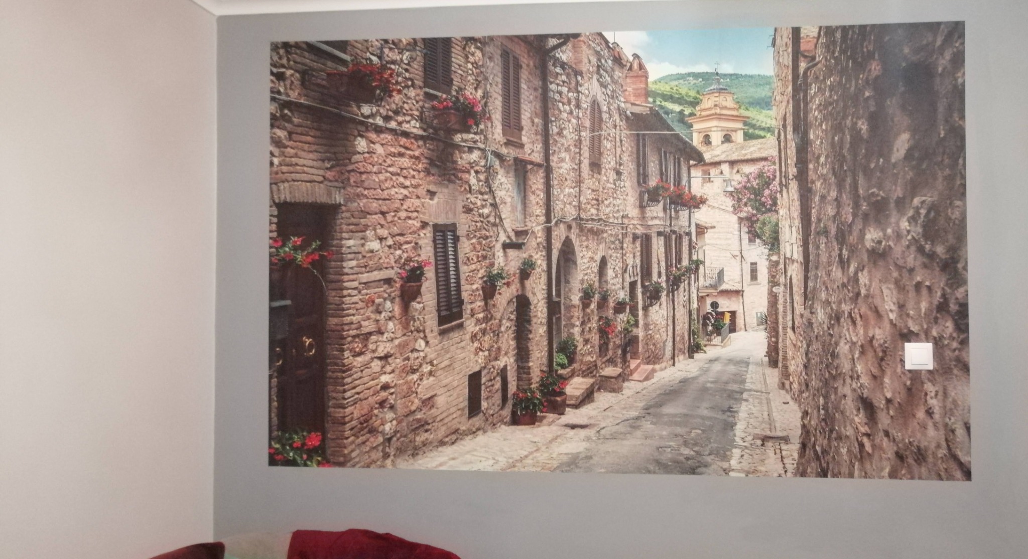 Fototapeta na ścianie przedstawiająca wąską uliczkę w toskańskim miasteczku z kamiennymi budynkami i kwiatami w doniczkach, z widocznym włącznikiem światła po prawej stronie.