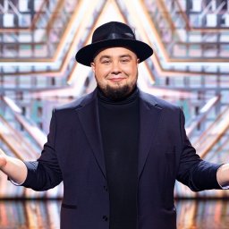 EVIVA LARTE MICHAŁ SKUBIDA - Mężczyzna w czarnym kapeluszu i granatowym garniturze, z brodą, uśmiechający się i prezentujący otwarte dłonie na tle geometrycznej dekoracji scenicznej.