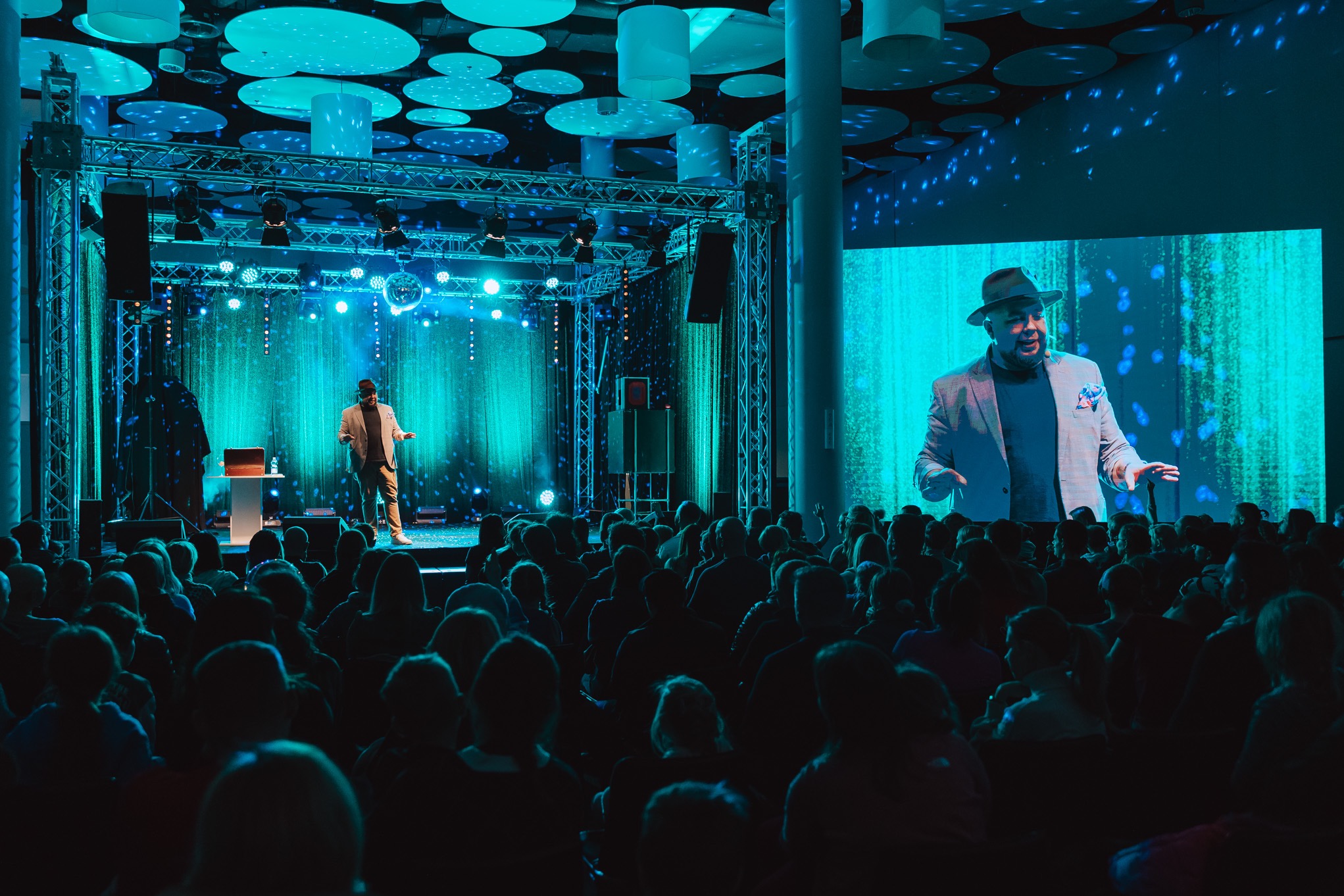 Scena z występem iluzjonisty przed publicznością, oświetlona niebieskimi światłami, z dużym ekranem projekcyjnym w tle.