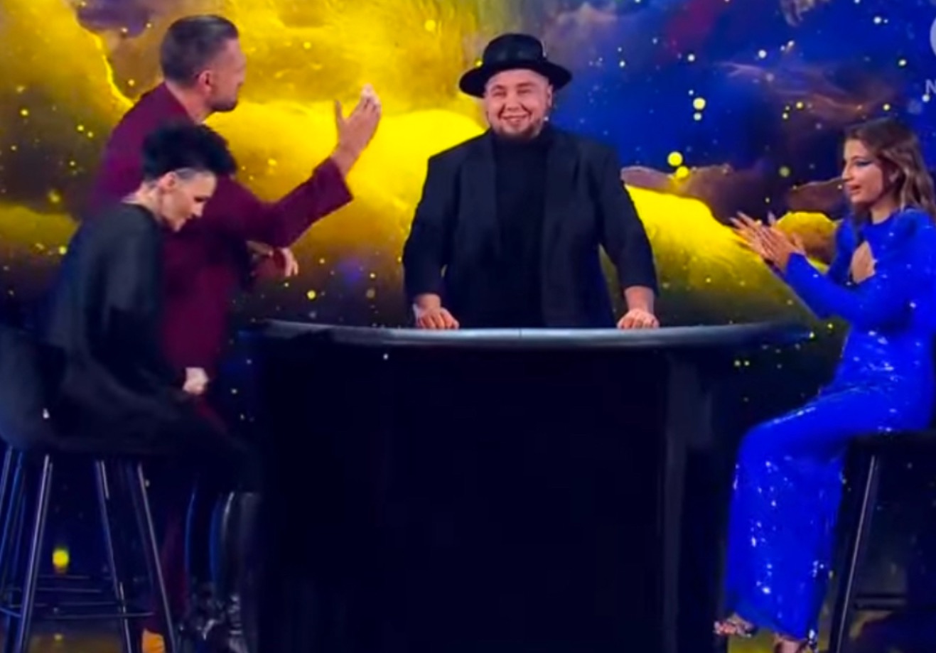 Iluzjonista w czarnym kapeluszu i marynarce prezentuje trik na czarnym stole, otoczony przez publiczność, z których jedna osoba ubrana jest w błyszczącą niebieską sukienkę, a tło stanowi...