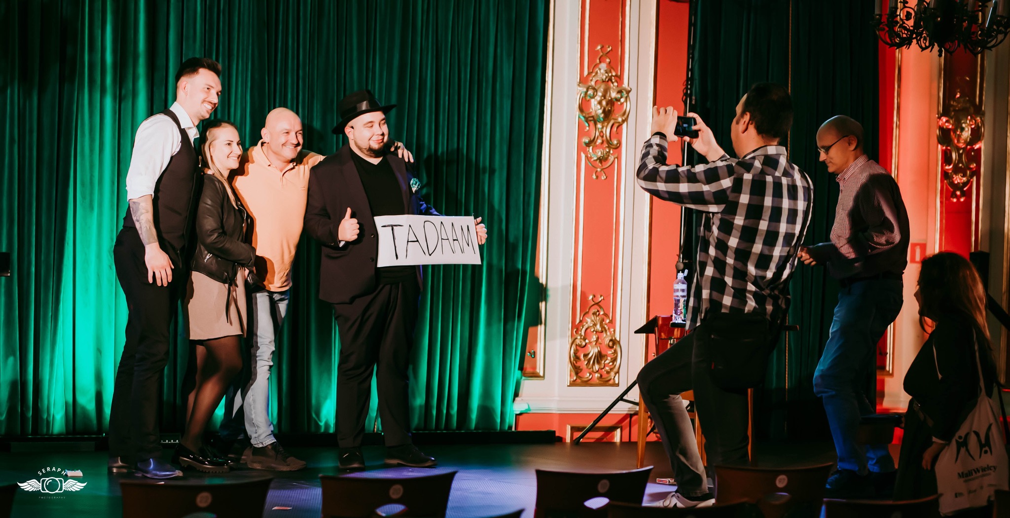 Iluzjonista w czarnym kapeluszu i garniturze, trzymający kartkę z napisem 'TADAAM', pozuje do zdjęcia z grupą osób na tle zielonej zasłony i dekoracyjnej ściany, podczas gdy inna osoba robi im...