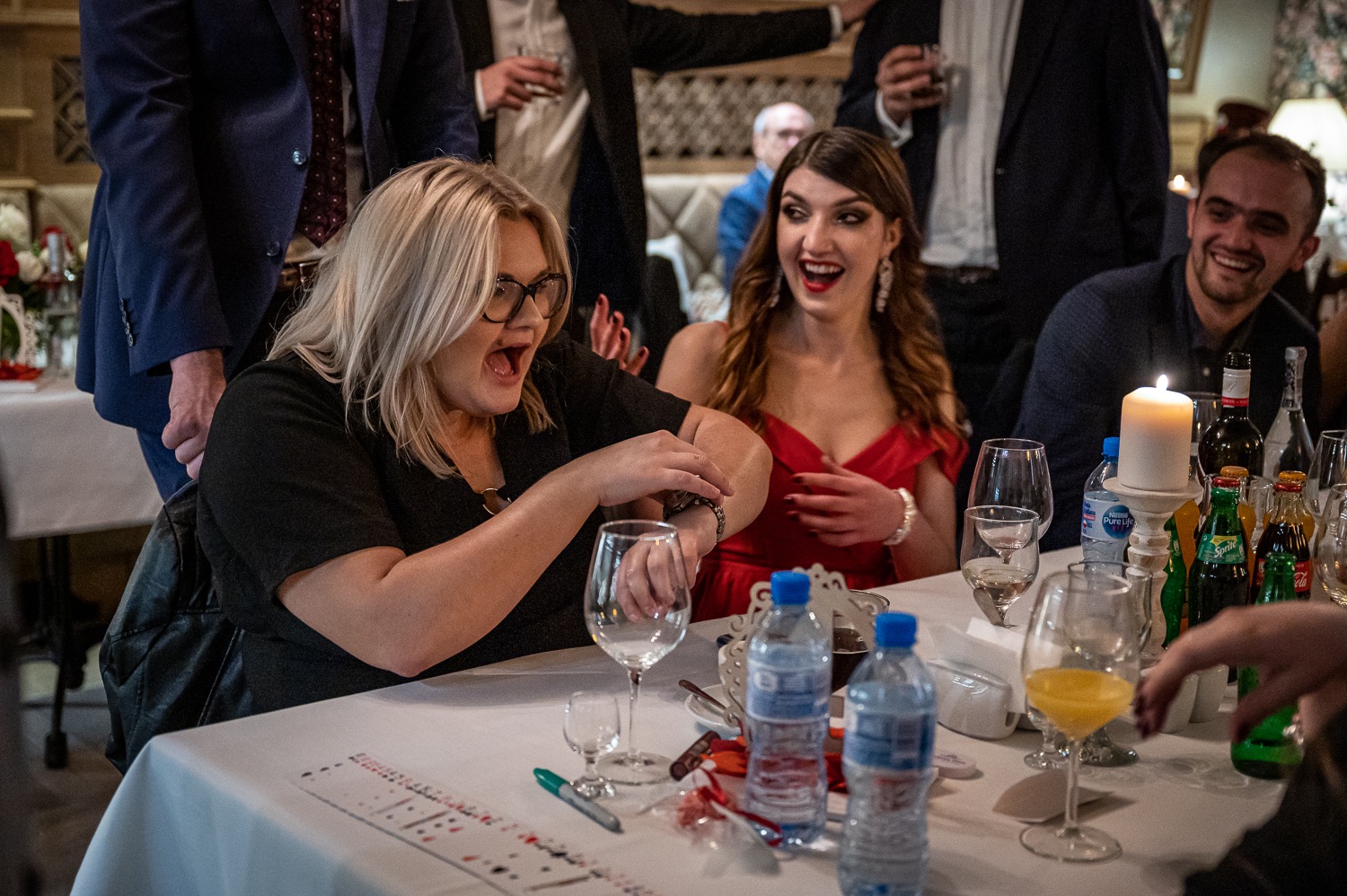 Kobieta w czarnej bluzce z otwartymi ustami patrzy na swój zegarek podczas pokazu iluzjonisty przy stole z kartami i publicznością w tle.