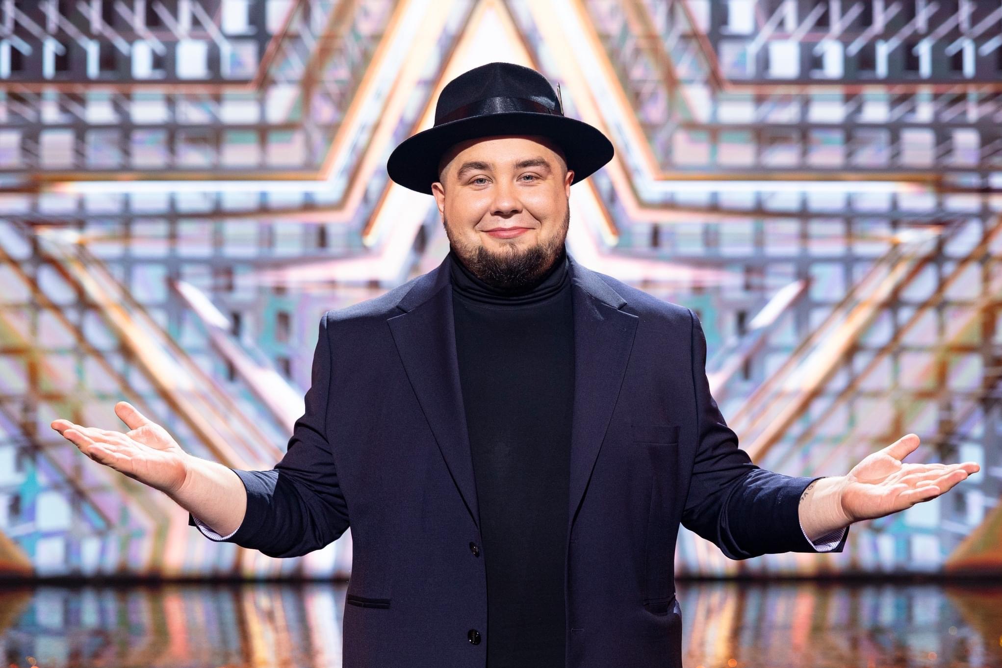Mężczyzna w czarnym kapeluszu i granatowym garniturze, z brodą, uśmiechający się i prezentujący otwarte dłonie na tle geometrycznej dekoracji scenicznej.