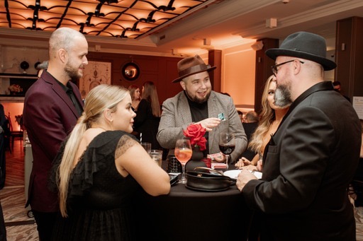 Iluzjonista w brązowym kapeluszu prezentuje kartę widzom podczas występu na przyjęciu, na stole kieliszki z drinkami i czerwona róża.