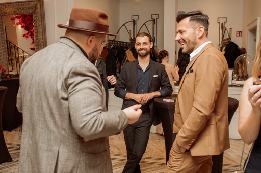 Mężczyzna w brązowym kapeluszu i tweedowej marynarce prezentuje trik karciany dwóm uśmiechniętym mężczyznom w garniturach na tle eleganckiego wnętrza.