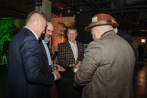 Mężczyźni w garniturach i kapeluszu obserwują pokaz iluzjonisty przy czarnym stoliku podczas imprezy firmowej, w tle zielona dekoracja.