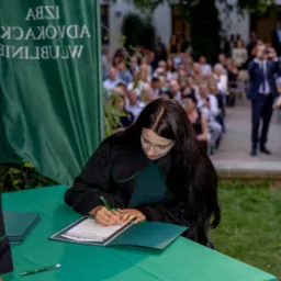 Kobieta w todze podpisuje dokument na tle widowni i zielonego baneru Izby Adwokackiej. Uroczystość wręczenia dyplomów.