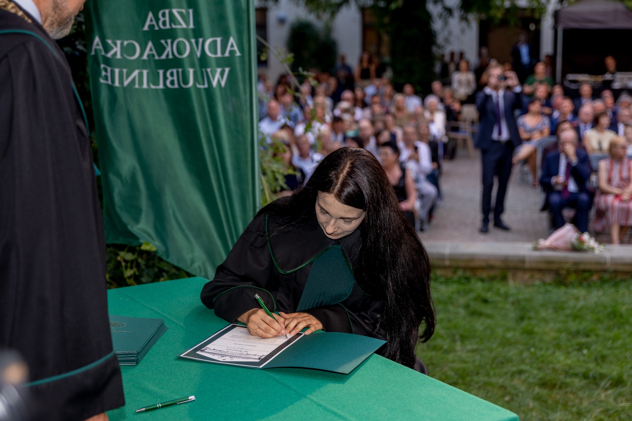 Kobieta w todze podpisuje dokument na tle widowni i zielonego baneru Izby Adwokackiej. Uroczystość wręczenia dyplomów.