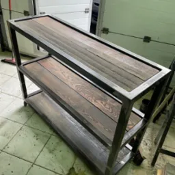 Metalowy regał na kółkach z trzema drewnianymi półkami o ciemnym wybarwieniu, widoczne ślady spawania, tło stanowią białe drzwi garażowe.