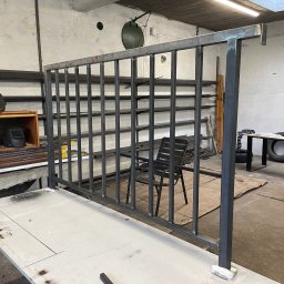 Michał Jerzewski - Szkielet metalowej konstrukcji, prawdopodobnie ogrodzenia lub balustrady, w warsztacie ślusarskim; widoczne spawane elementy, maski spawalnicze na półce, oraz zarys planu na stole.