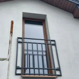 Zewnętrzna, metalowa balustrada okienna z drewnianymi elementami mocowana do ściany budynku z widocznym ociepleniem w szczelinie obok okna.