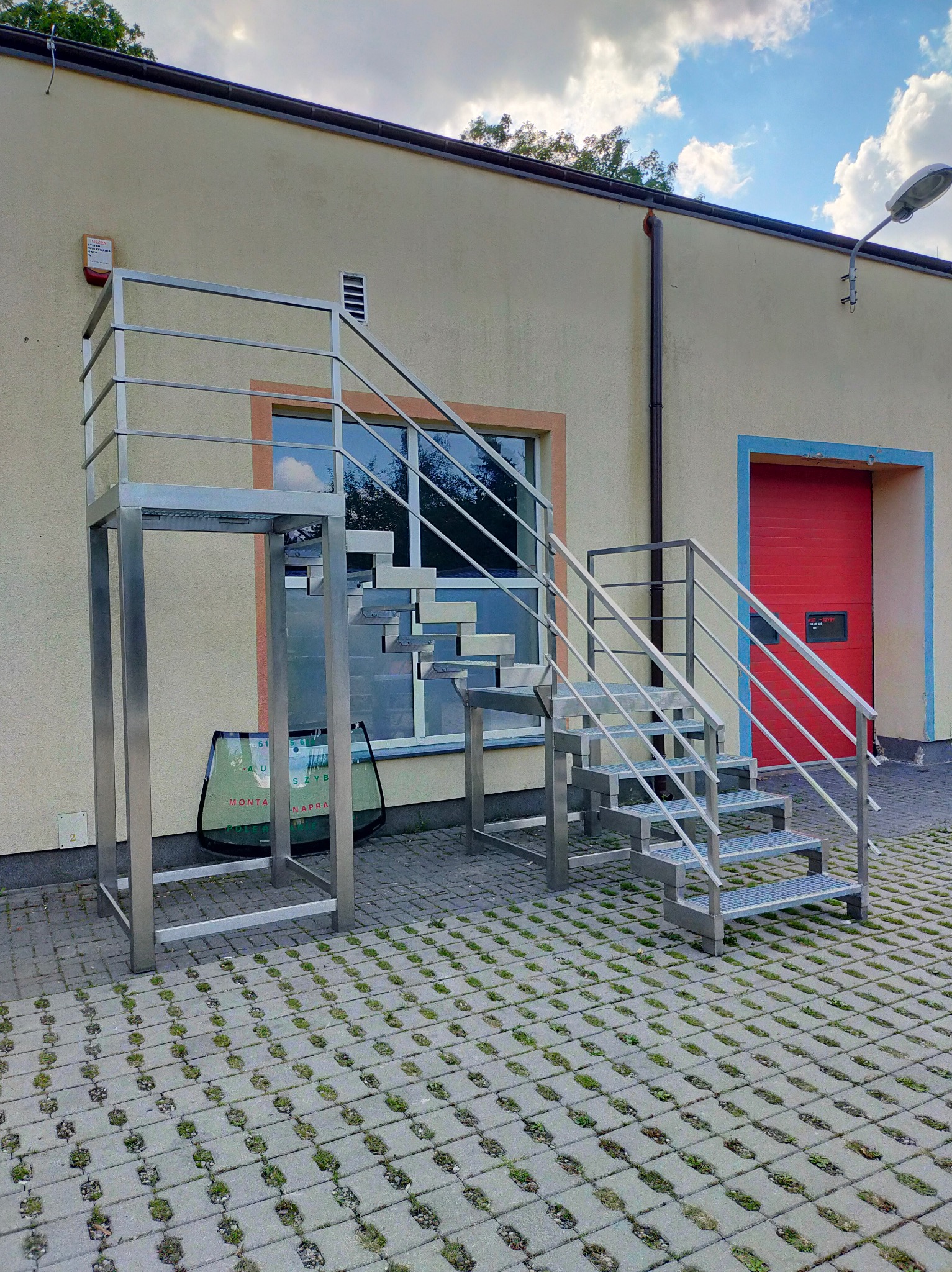 Zewnętrzne schody metalowe z platformą, prowadzące do wejścia na budynek, z widoczną szybą samochodową w tle.