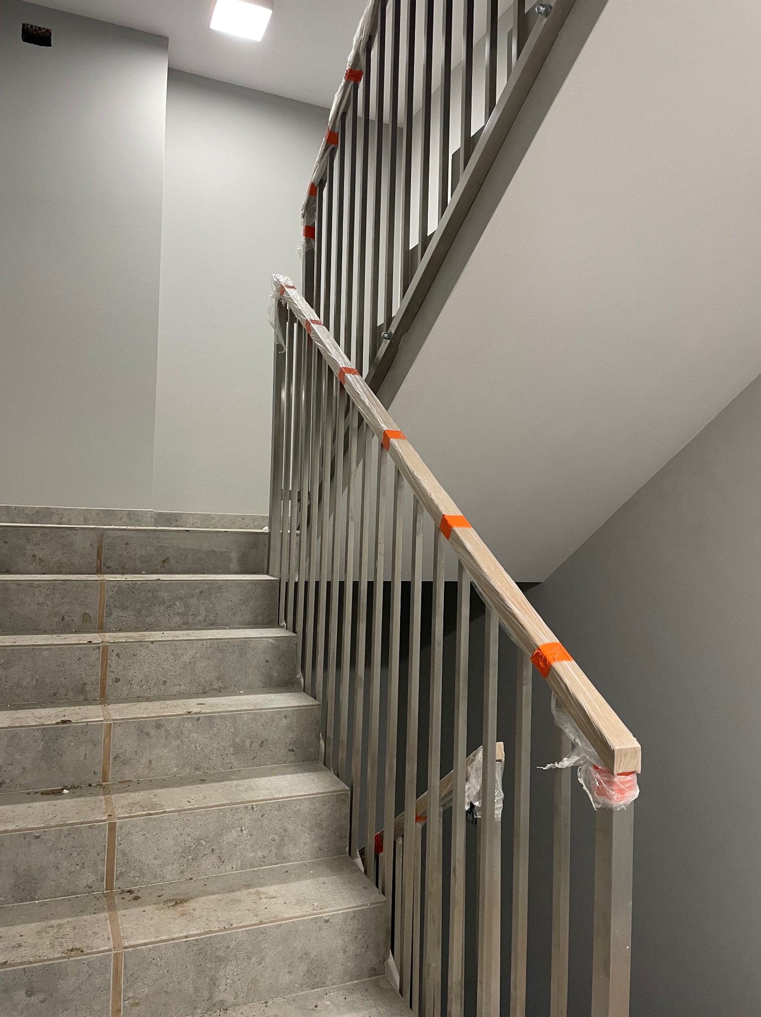 Schody wewnątrz budynku z metalową balustradą, zabezpieczoną folią i pomarańczową taśmą, widoczne niedociągnięcia w wykończeniu schodów.