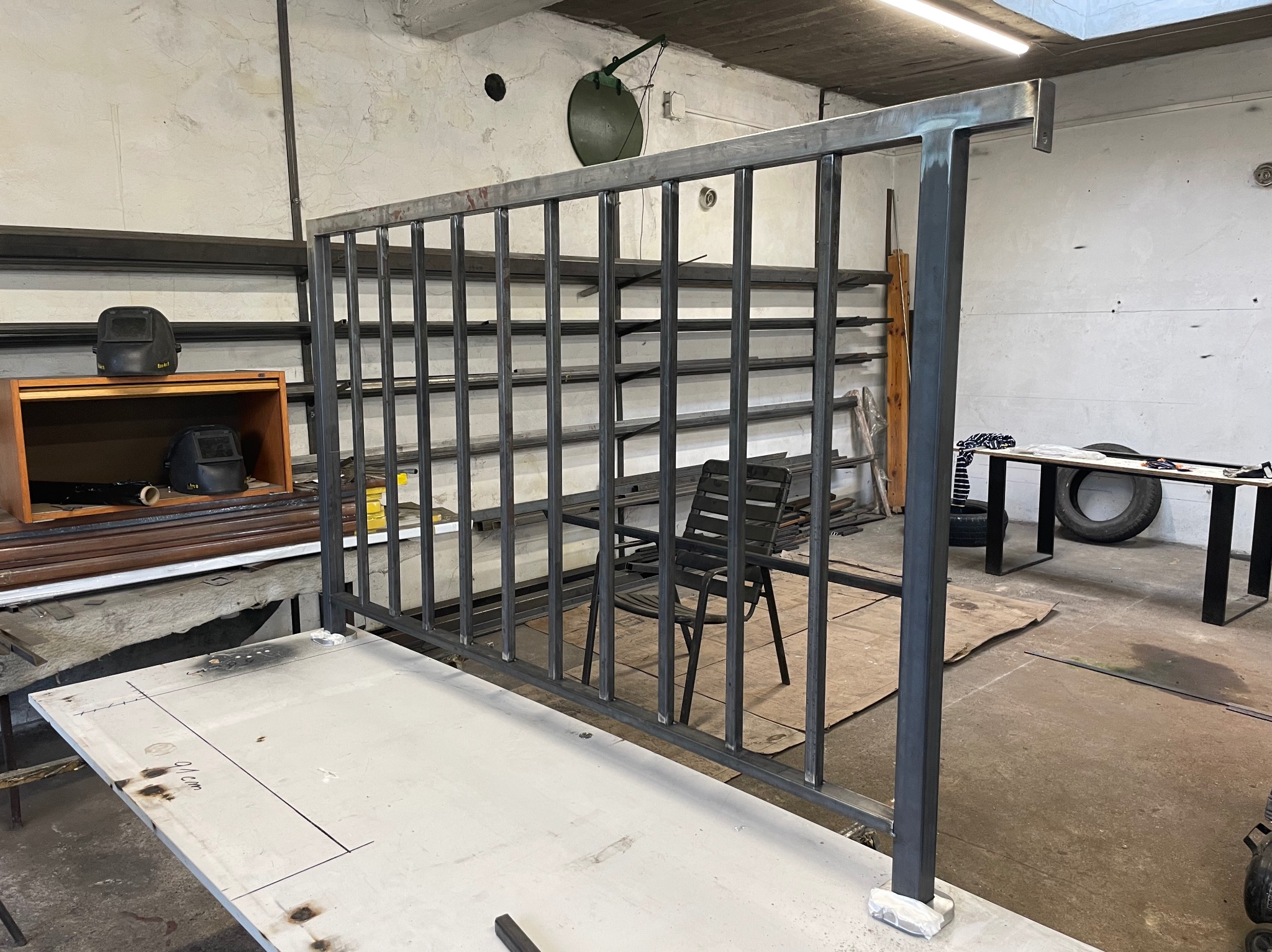 Szkielet metalowej konstrukcji, prawdopodobnie ogrodzenia lub balustrady, w warsztacie ślusarskim; widoczne spawane elementy, maski spawalnicze na półce, oraz zarys planu na stole.