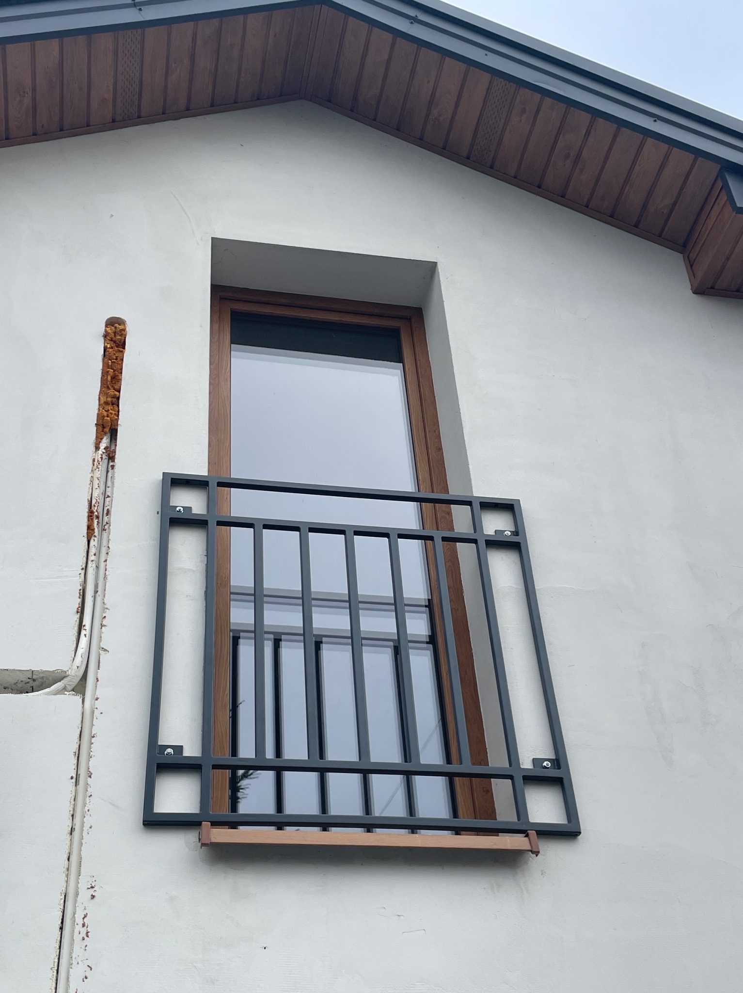 Zewnętrzna, metalowa balustrada okienna z drewnianymi elementami mocowana do ściany budynku z widocznym ociepleniem w szczelinie obok okna.