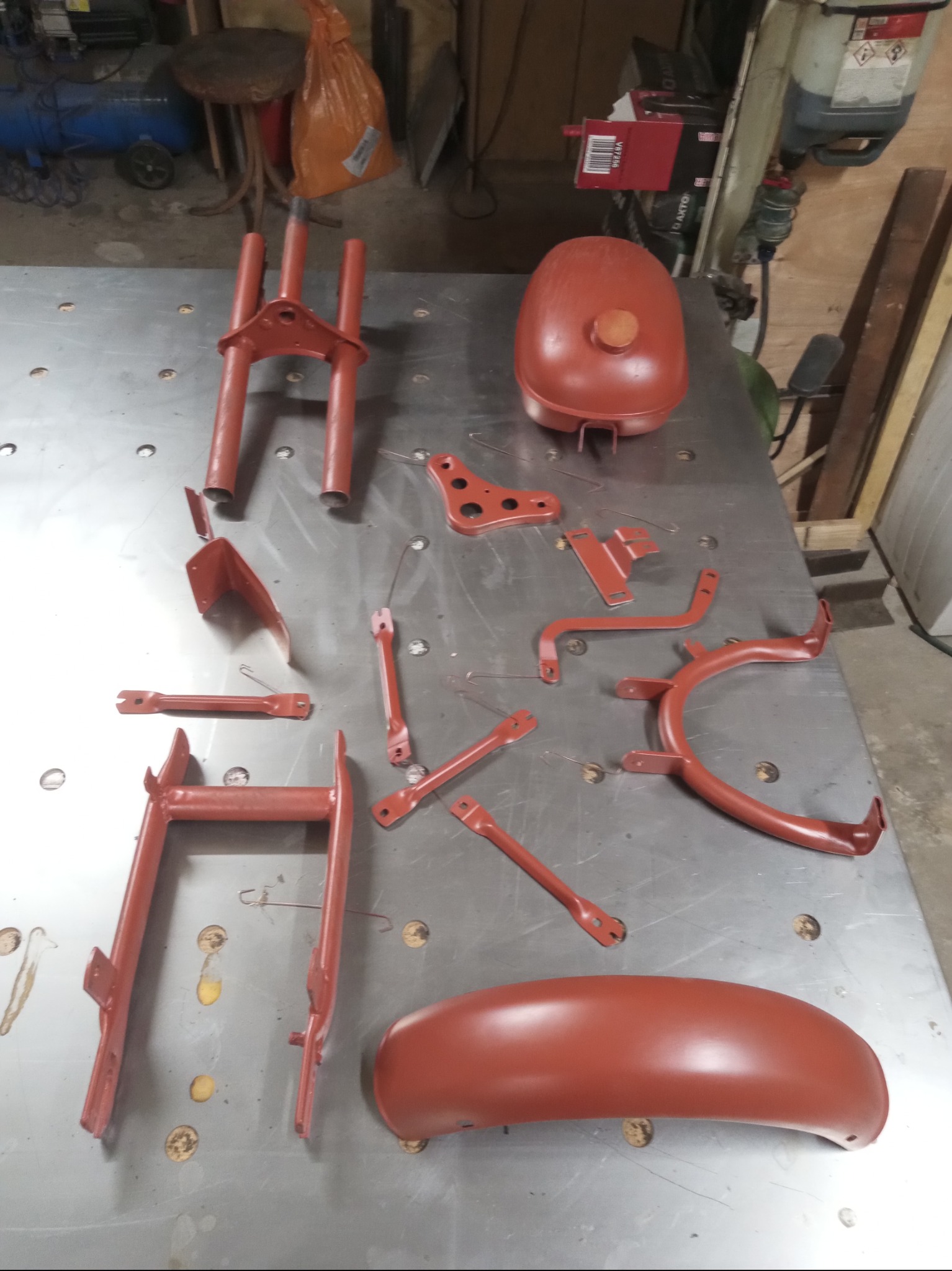 Pomalowane na czerwono elementy motocykla leżące na metalowym stole warsztatowym. Widoczne błotniki, zbiornik paliwa i inne drobne części.