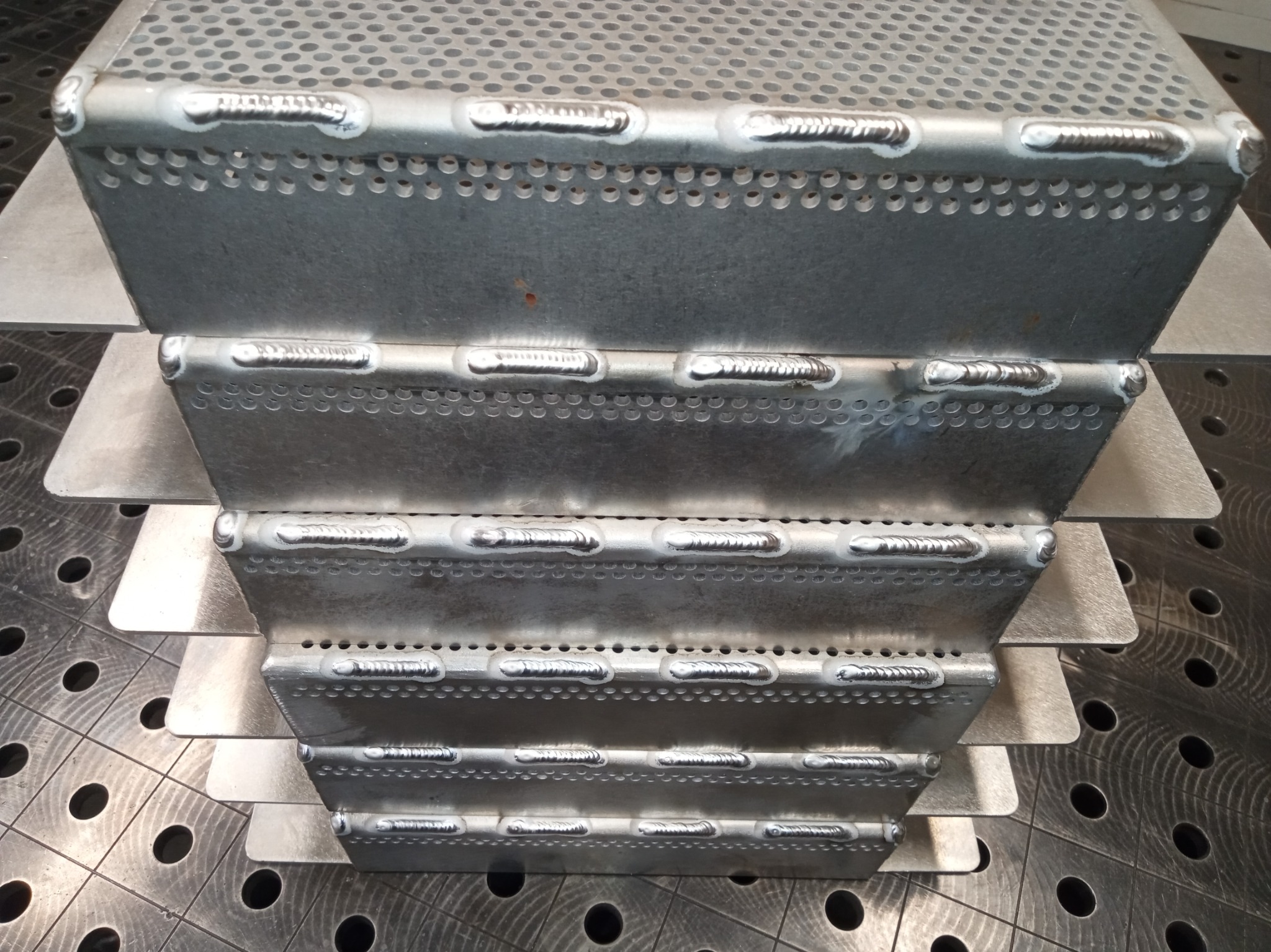 Metalowa konstrukcja schodkowa z perforowanej blachy, połączona spawami. Widoczne regularne spoiny i otwory. Tło stanowi metalowy stół spawalniczy z otworami.