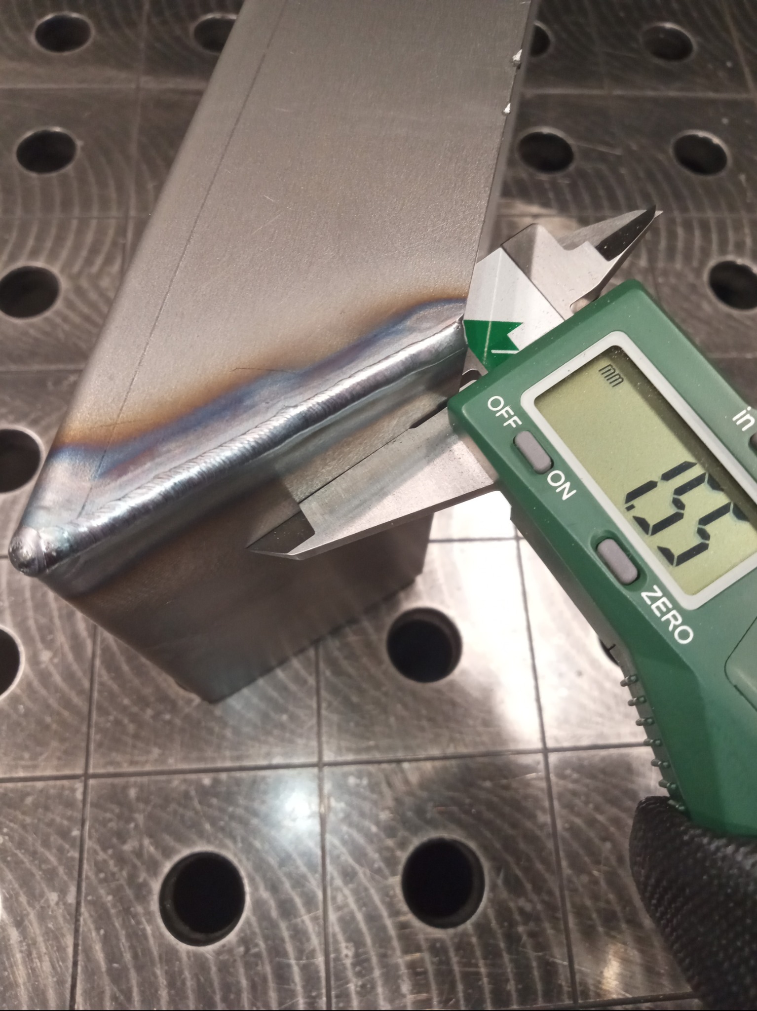 Pomiar spoiny spawalniczej suwmiarką elektroniczną na perforowanym stole spawalniczym. Widoczna precyzja wykonania i kontrola jakości.