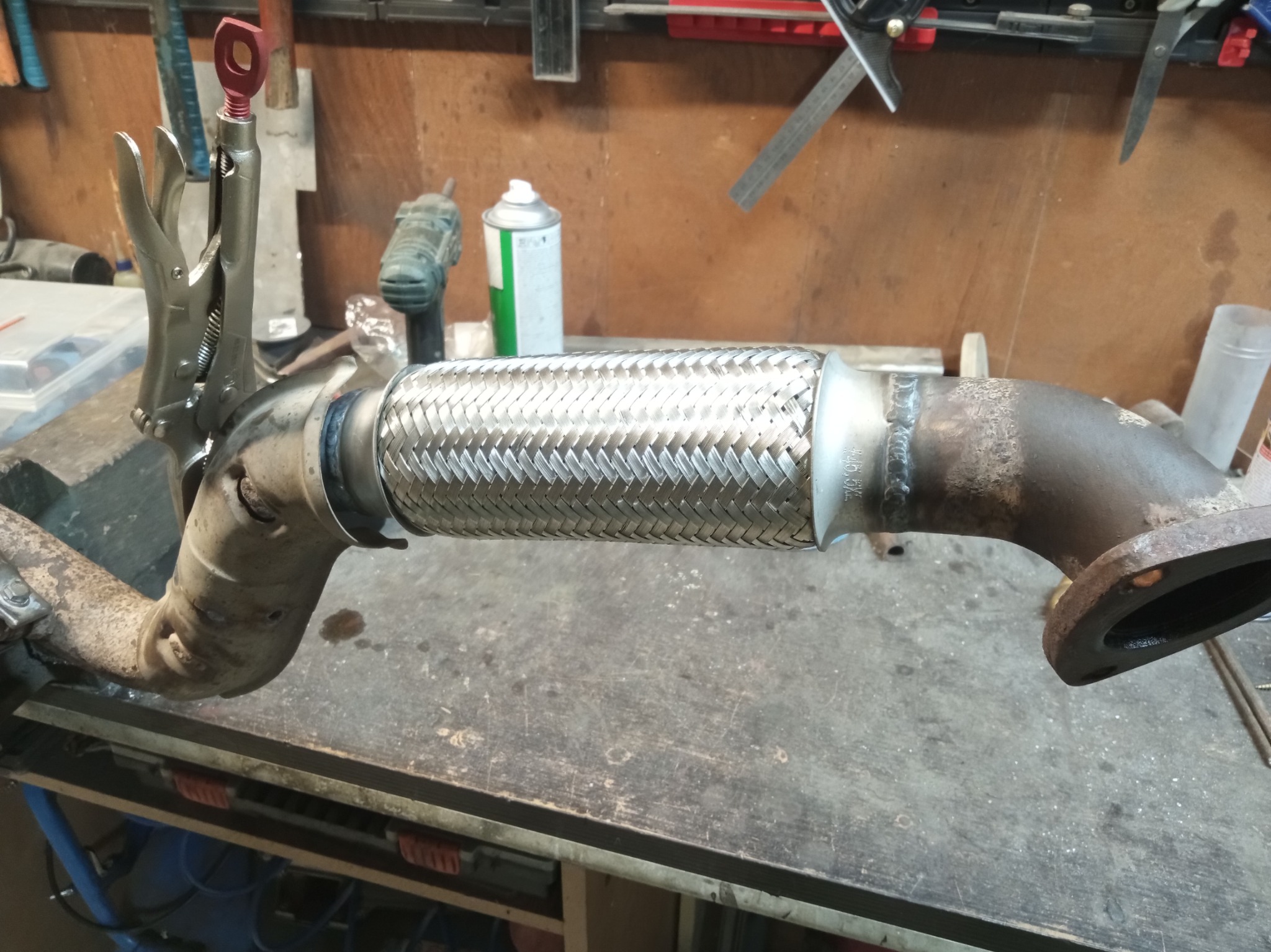 Spawany element układu wydechowego z metalowym oplotem, zamocowany na warsztatowym stole. Widoczne narzędzia i akcesoria spawalnicze w tle.