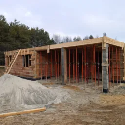 Budowa parterowego domu jednorodzinnego w stanie surowym otwartym: widoczne ściany z cegły, szalunki, podpory stropowe w kolorze pomarańczowym oraz pryzma piasku na pierwszym planie.