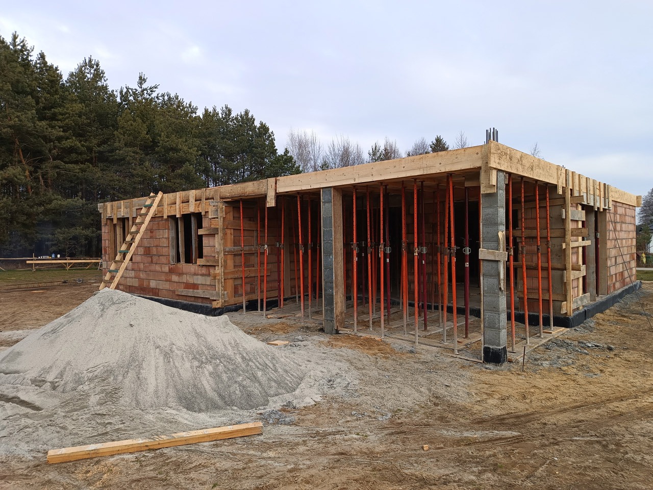 Budowa parterowego domu jednorodzinnego w stanie surowym otwartym: widoczne ściany z cegły, szalunki, podpory stropowe w kolorze pomarańczowym oraz pryzma piasku na pierwszym planie.