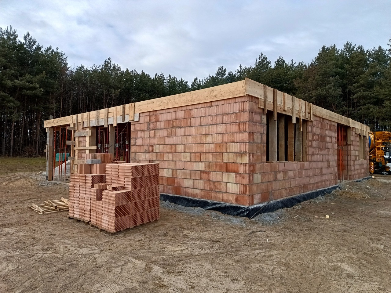 Budowa domu jednorodzinnego w stanie surowym otwartym, z widocznymi bloczkami ceramicznymi, drewnianymi elementami konstrukcyjnymi i stosem bloczków obok, na tle lasu.