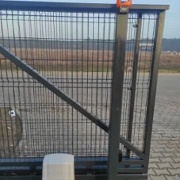 Przesuwna brama z siatki z zamontowanym napędem Roger Technology i pomarańczową lampą ostrzegawczą, widoczna na tle pola i lasu.