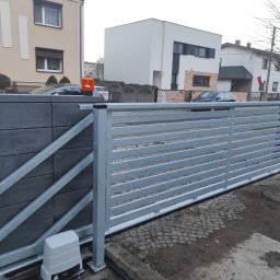 M-tech Michał Śliwczyński - Nowoczesna, przesuwna brama wjazdowa z szarego metalu, z widocznym napędem elektrycznym i słupkiem z szarej kostki, na tle zabudowy miejskiej.