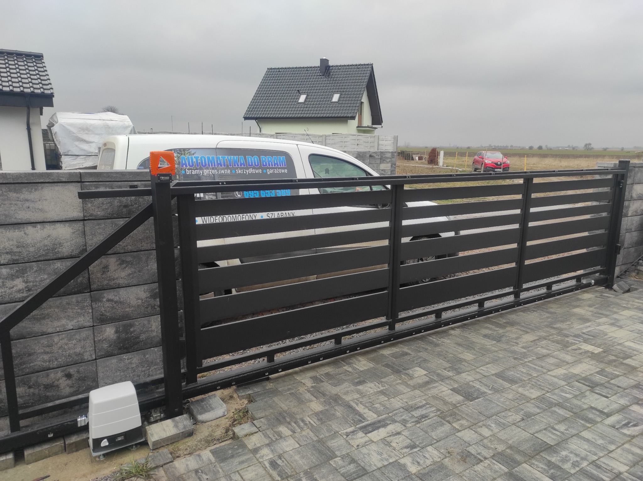 Zakończona realizacja: Czarna, przesuwna brama wjazdowa z automatyką, na posesji z szarej kostki brukowej, w tle budynek i samochód firmy z reklamą automatyki do bram.
