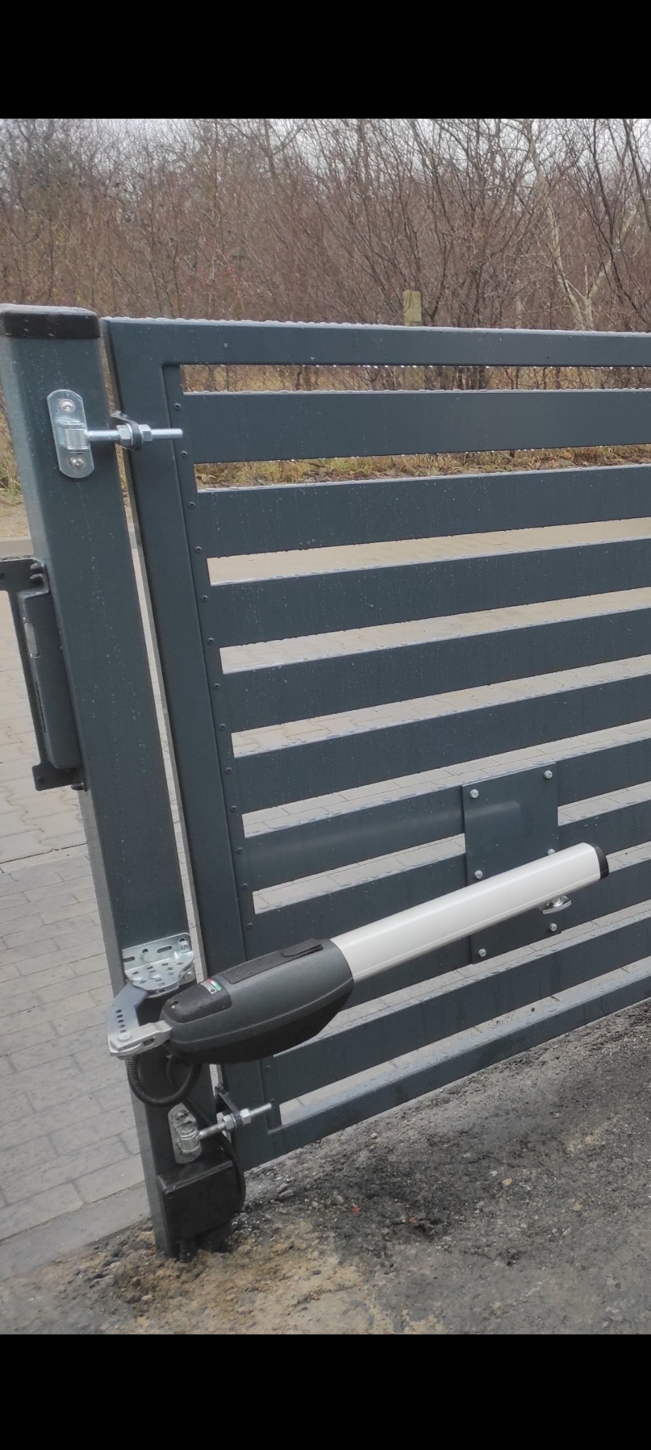 Szara brama wjazdowa z automatyką, widoczne elementy mocujące i siłownik, tło stanowią drzewa bez liści.