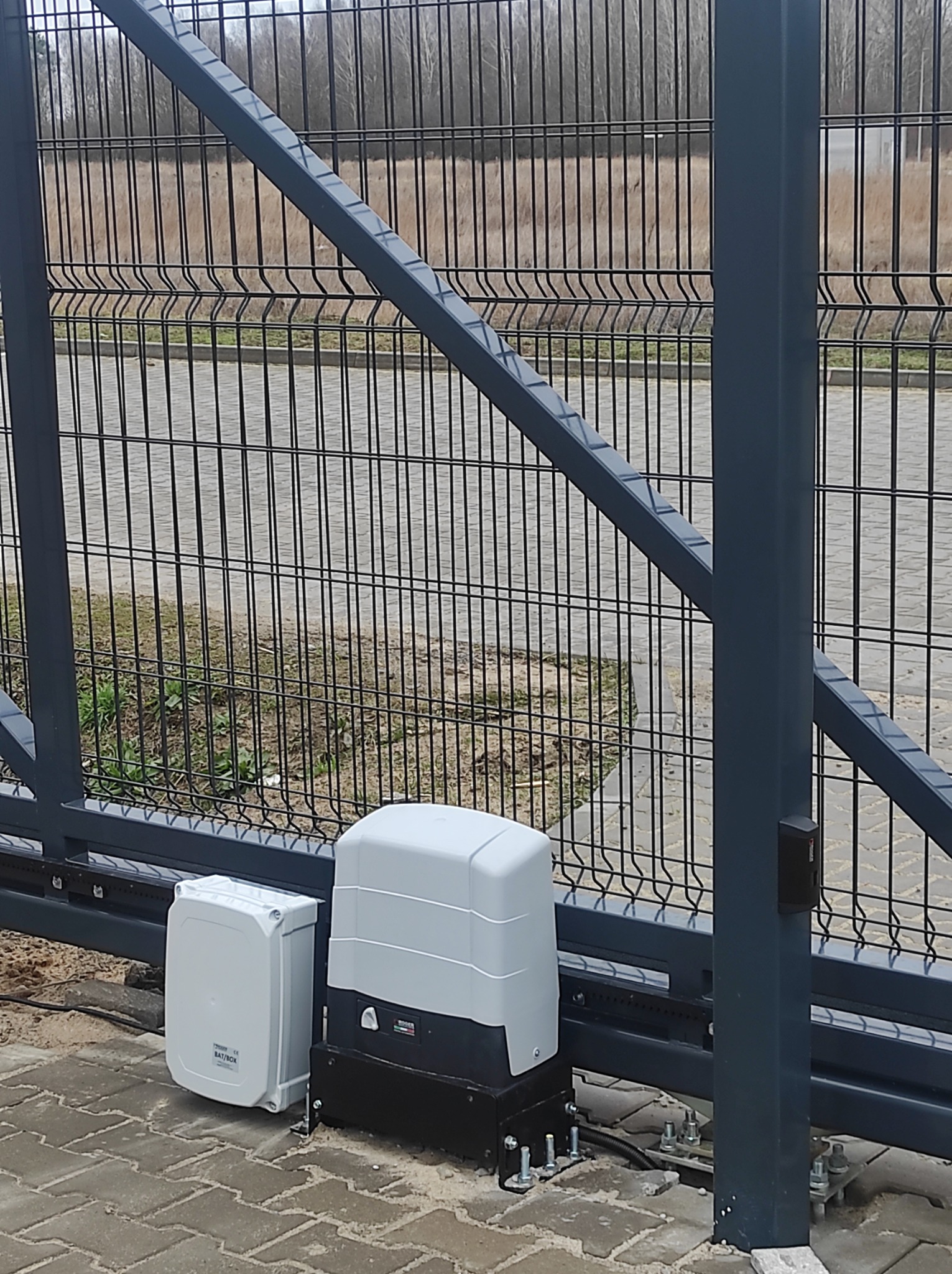 Szara, przesuwna brama z siatki z zamontowanym napędem i skrzynką sterującą na kostce brukowej.