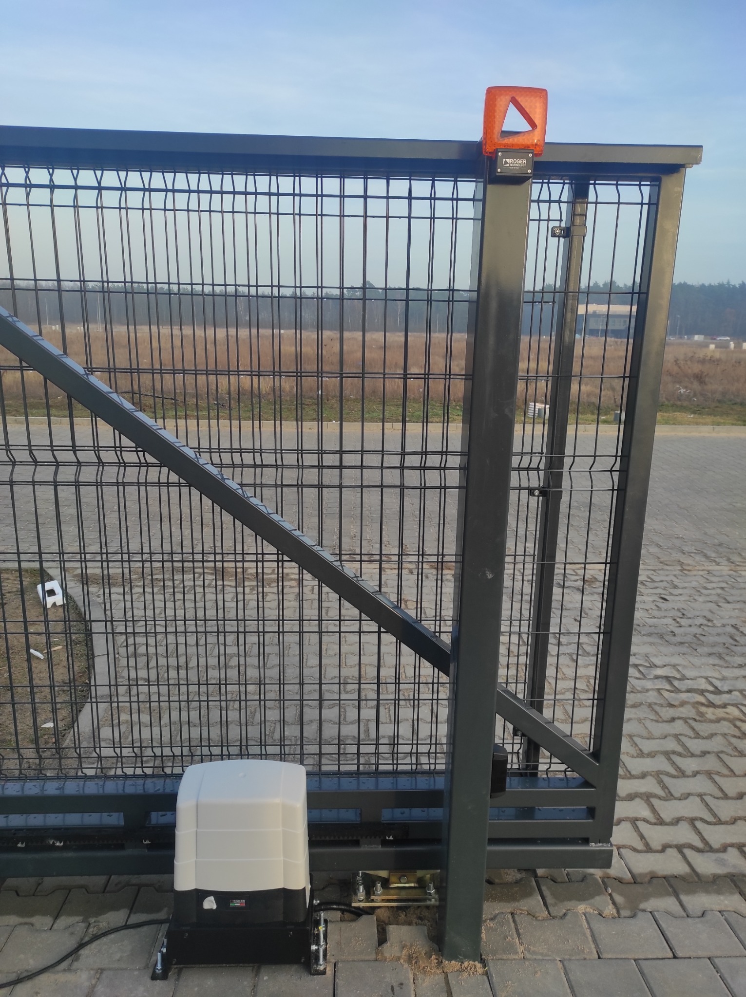 Przesuwna brama z siatki z zamontowanym napędem Roger Technology i pomarańczową lampą ostrzegawczą, widoczna na tle pola i lasu.