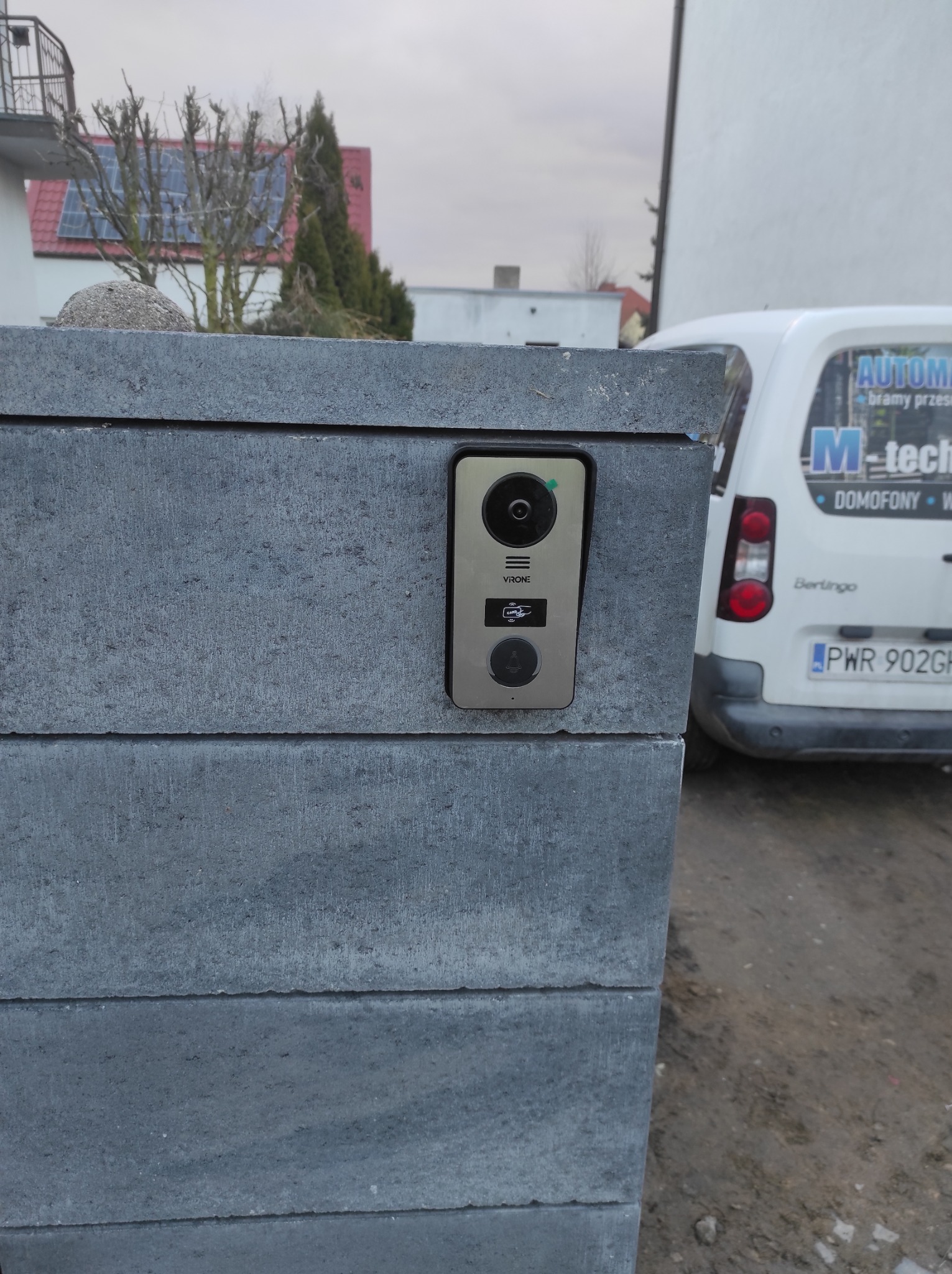 Zamontowany wideofon Vione na murowanym słupku ogrodzenia, z widocznym samochodem dostawczym firmy instalacyjnej w tle.