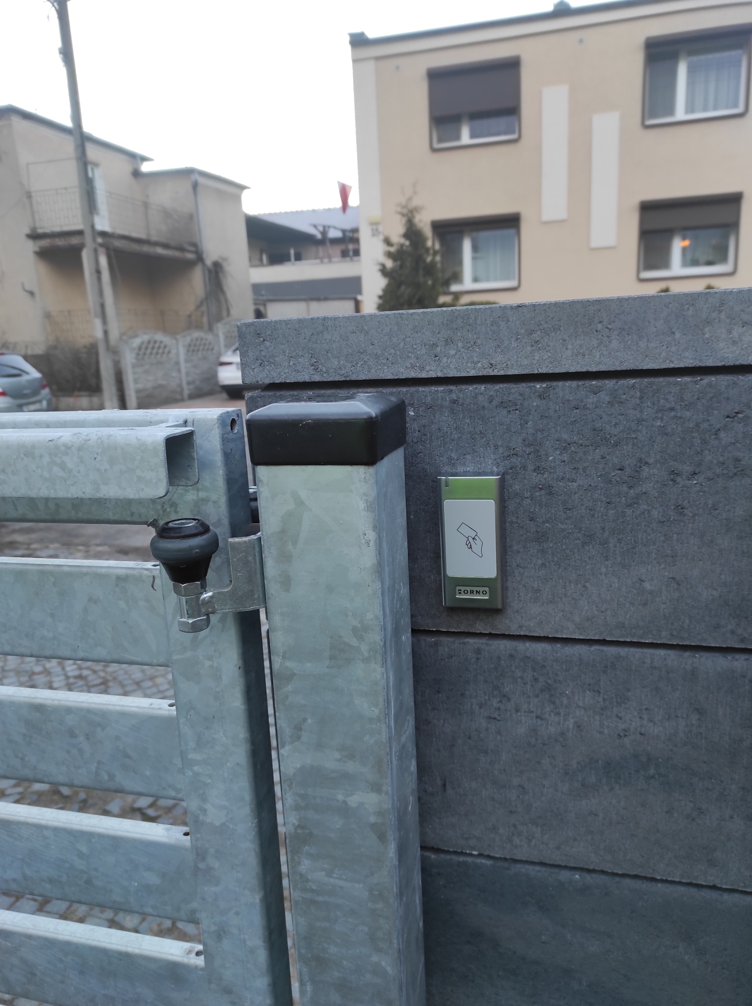 Szara brama wjazdowa z czytnikiem kart zbliżeniowych ZORNO zamontowanym na betonowym słupku.