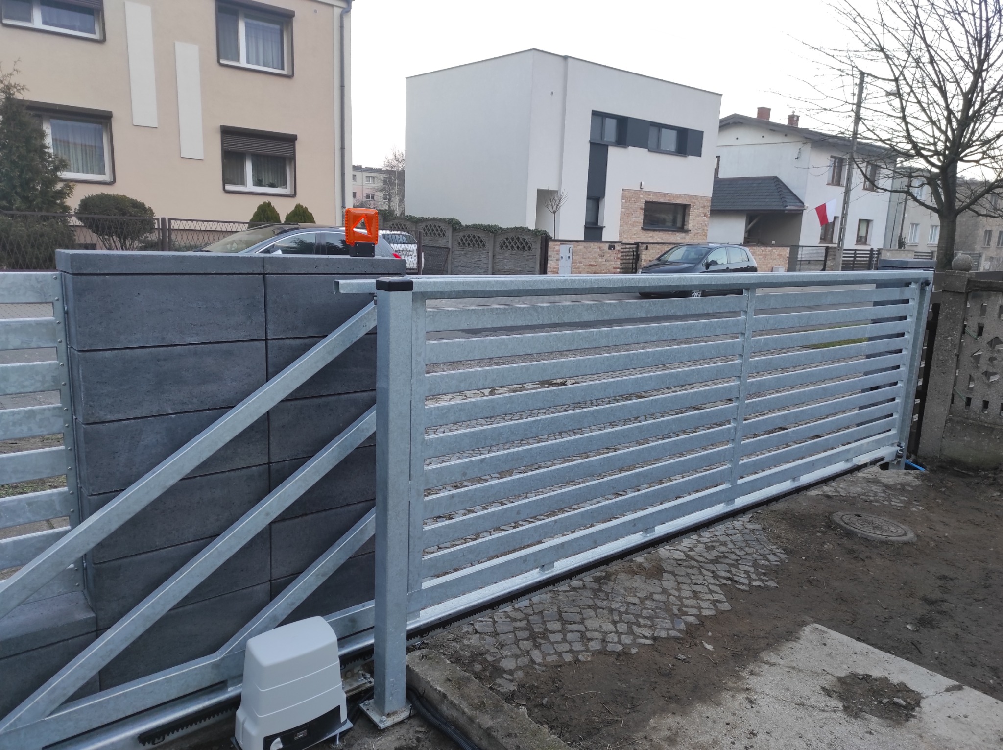 Nowoczesna, przesuwna brama wjazdowa z szarego metalu, z widocznym napędem elektrycznym i słupkiem z szarej kostki, na tle zabudowy miejskiej.