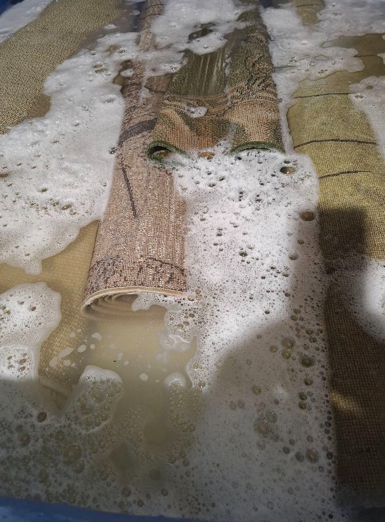 Fragment prania kilku dywanów w wannie, widoczna piana i brudna woda.