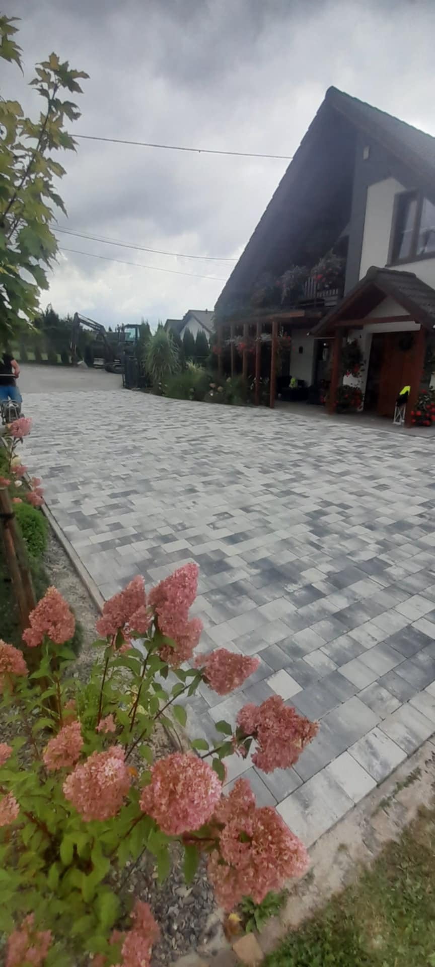 Szara kostka brukowa ułożona na podjeździe przed domem z kwitnącymi, różowymi hortensjami na pierwszym planie i koparką w tle.