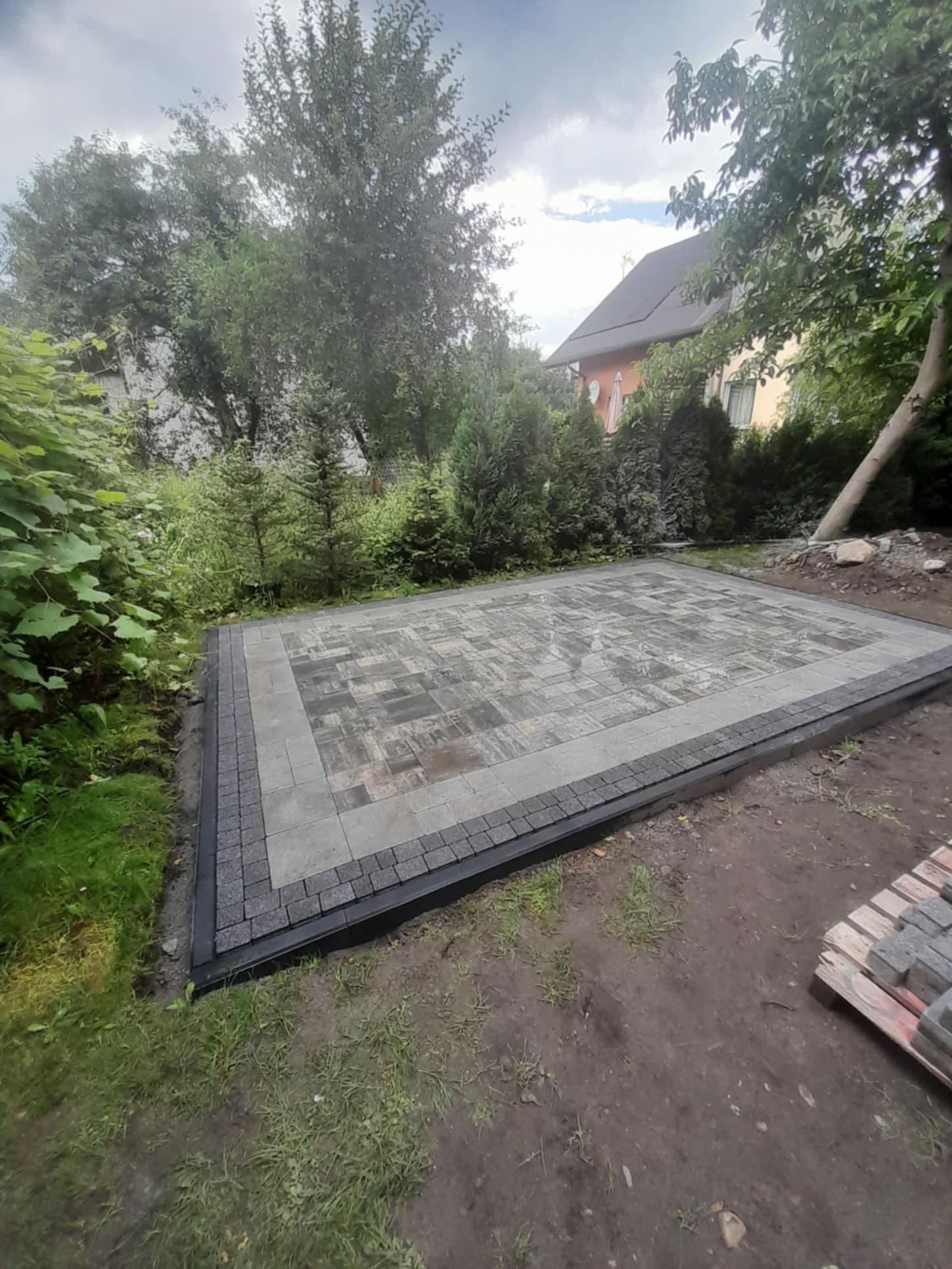 Nowo ułożony plac z kostki brukowej w odcieniach szarości z obramowaniem z ciemniejszej kostki, widoczne przygotowane palety kostki, tło z zieleni i fragment budynku.