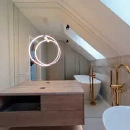 Nowoczesna łazienka na poddaszu z dużym lustrem, drewnianą szafką pod umywalką i złotą armaturą. Widoczne skosy i okno dachowe.