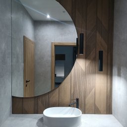 IRON GLASS SPÓŁKA Z OGRANICZONĄ ODPOWIEDZIALNOŚCIĄ - Nowoczesna łazienka z owalnym lustrem, drewnianą ścianą z geometrycznym wzorem i minimalistyczną umywalką nablatową. Widoczne drzwi z drewnianą ościeżnicą.