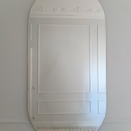 IRON GLASS SPÓŁKA Z OGRANICZONĄ ODPOWIEDZIALNOŚCIĄ - Owalne lustro z dekoracyjnymi listwami i ornamentami, zawieszone na jasnej ścianie. Minimalistyczny design z subtelnymi zdobieniami.