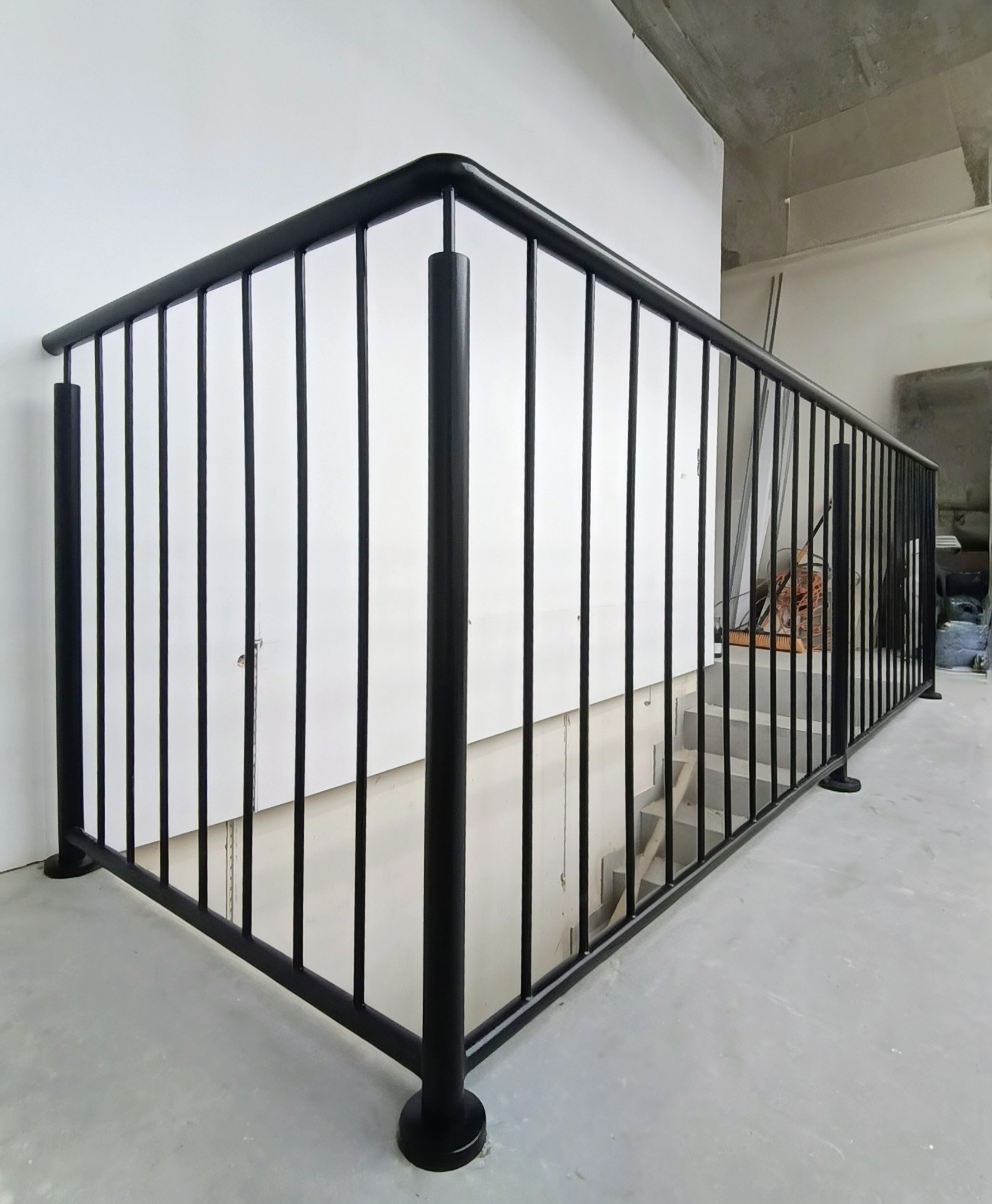 Nowoczesna, czarna balustrada schodowa z metalowymi prętami, zamontowana w surowym wnętrzu z betonowym sufitem. Minimalistyczny design, widok na schody.