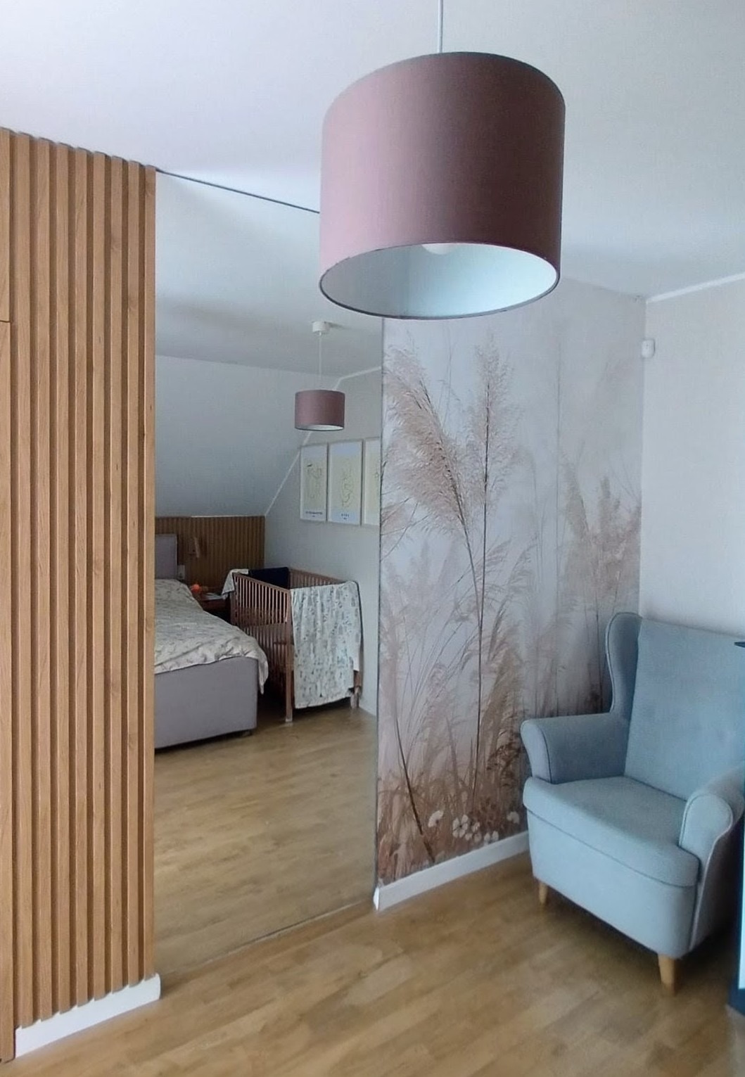 Szklana ściana działowa z drewnianymi lamelami oddziela pokój dziecięcy od salonu. Na ścianie fototapeta z motywem trawy pampasowej i błękitny fotel.