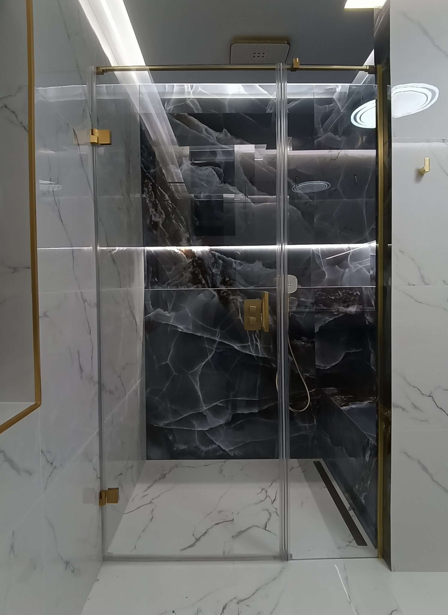 Nowoczesna kabina prysznicowa z przezroczystego szkła, złote okucia, marmurowe płytki w odcieniach bieli i szarości. Eleganckie wykończenie łazienki.